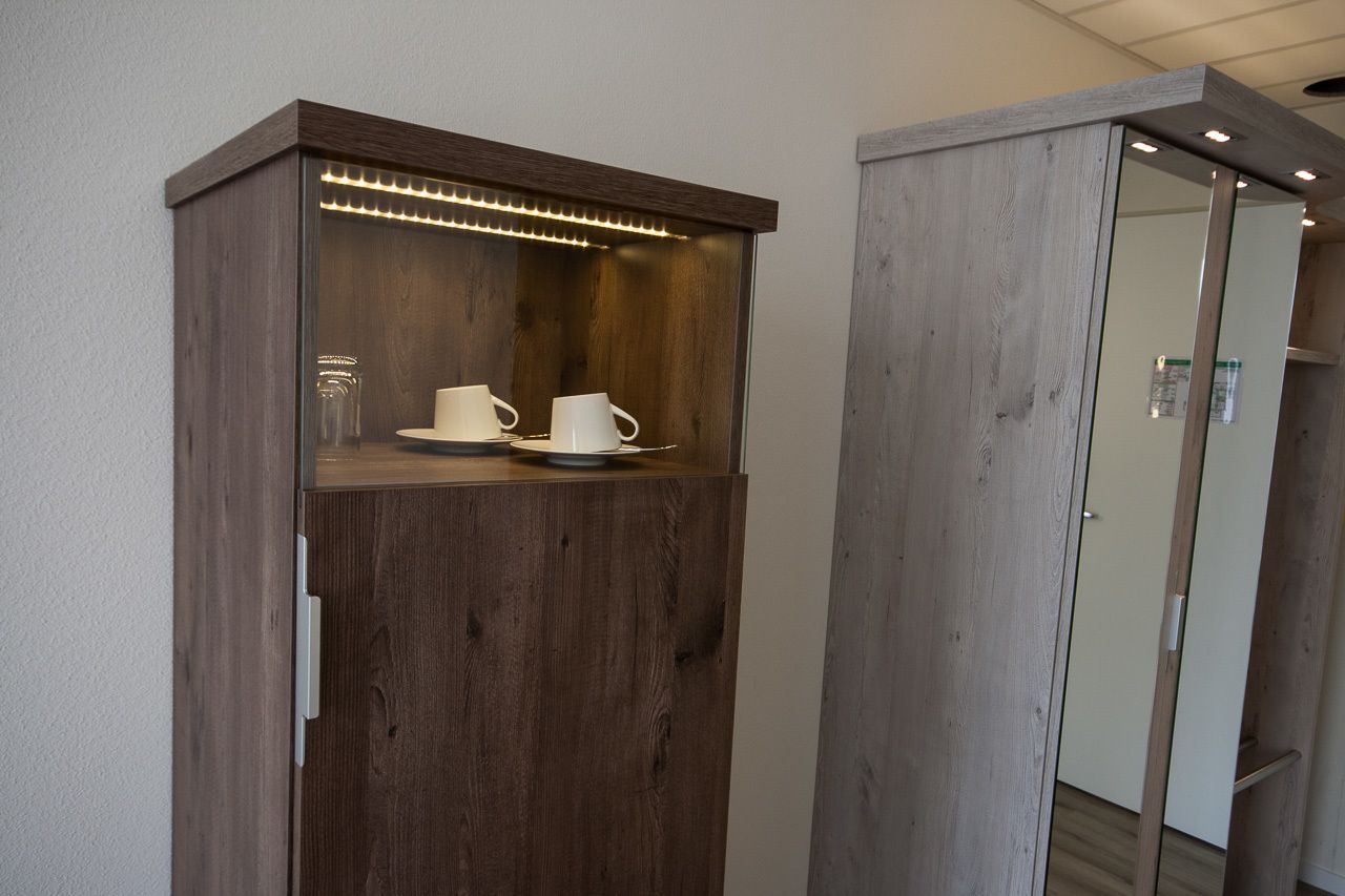 A wooden cabinet with a glass door, containing a white cup and saucer, and a glass jar.