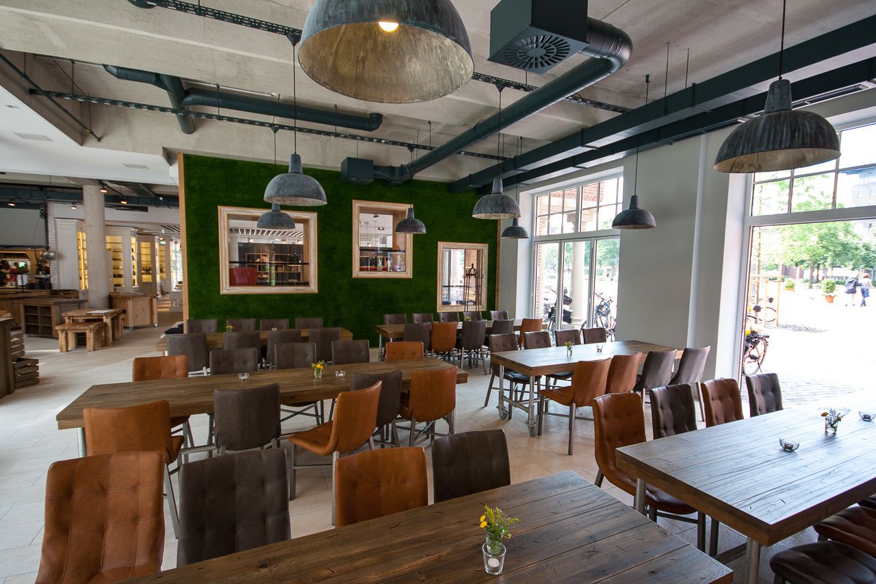 Un café amplio con mesas y sillas de madera, grandes lámparas colgantes y una pared verde con cuadros enmarcados.