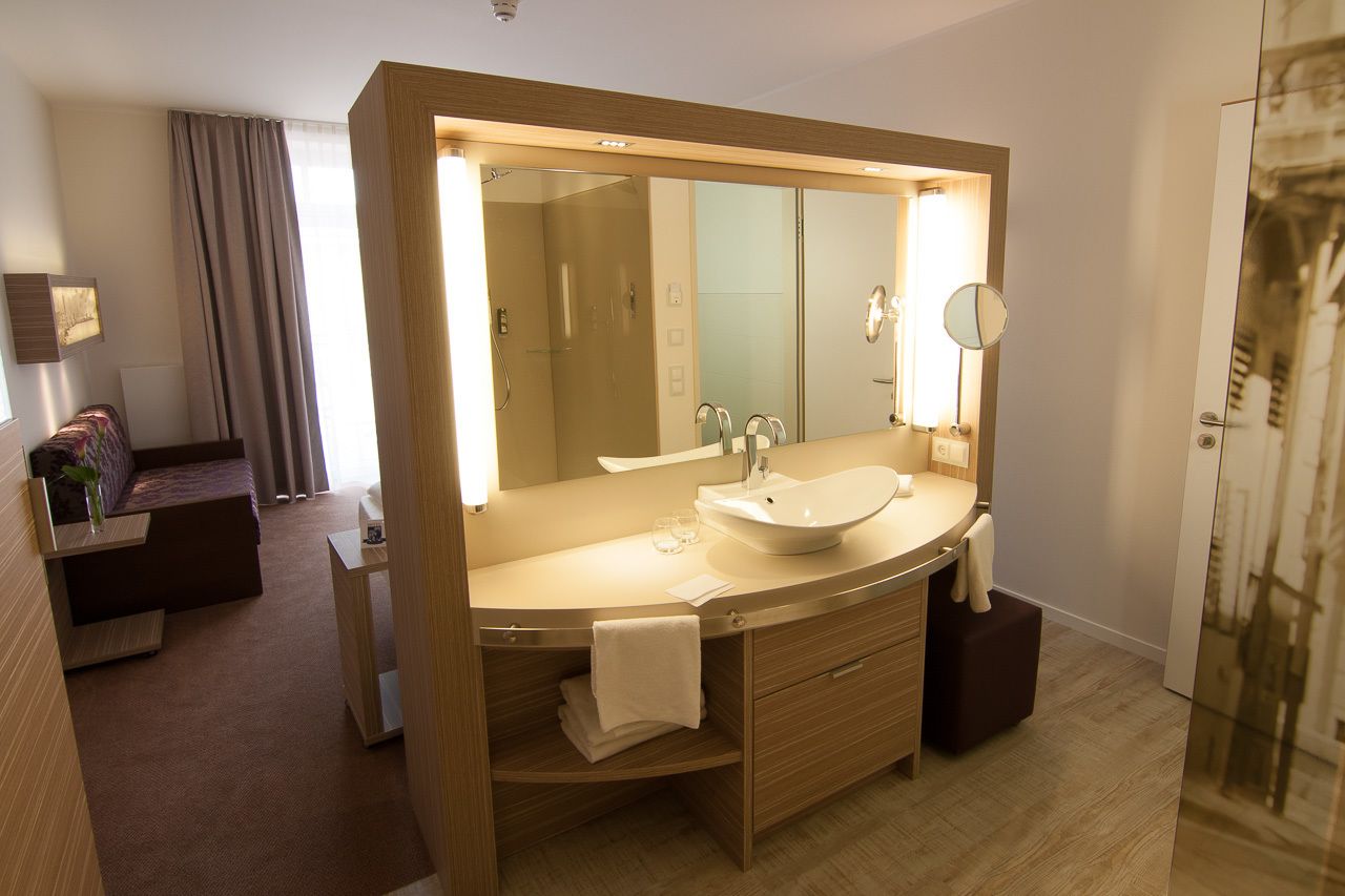 Een moderne badkamer met een grote spiegel, een wastafel en een douche. Er is een wastafelmeubel met een handdoekrek en een klein tafeltje met een vaas. De kamer heeft een houten vloer en een groot raam met gordijnen.