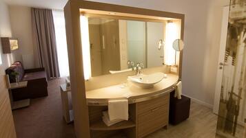 Een moderne badkamer met een grote spiegel, een wastafel en een douche. Er is een wastafelmeubel met een handdoekrek en een klein tafeltje met een vaas. De kamer heeft een houten vloer en een groot raam met gordijnen.