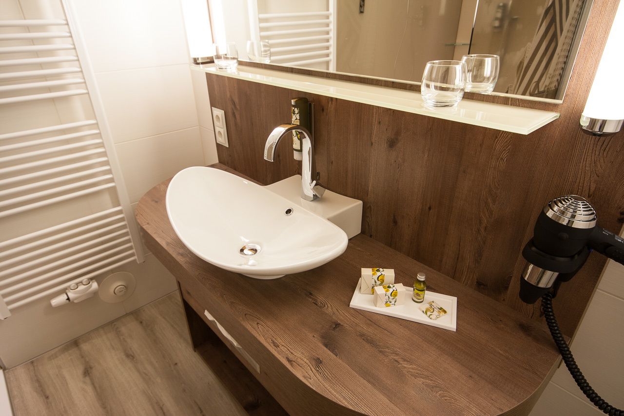 A bathroom with a white sink, wooden vanity, and a mirror above the sink. There is a towel rack to the left and a hairdryer on the right.