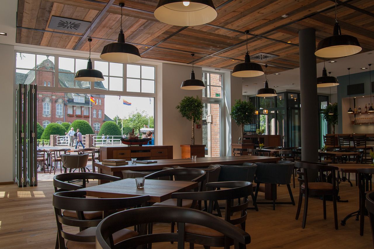 A spacious cafe with wooden tables and chairs, large windows offering a view of a red brick building and greenery outside.