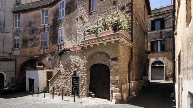 Une ruelle étroite pavée, bordée d’un bâtiment en pierre avec une porte noire et un petit escalier menant à un étage supérieur.
