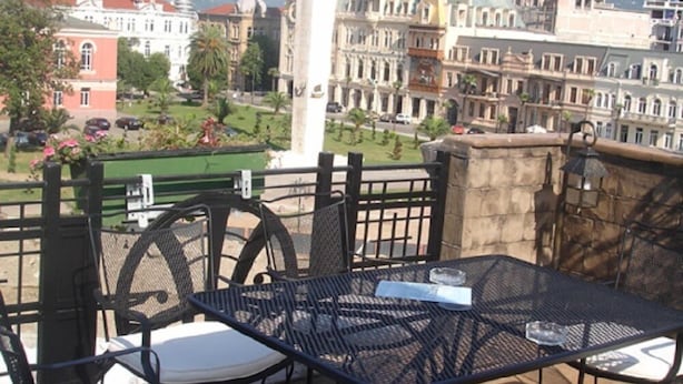 A terrace with outdoor seating, overlooking a cityscape with historic buildings and a statue. - Hotel Brighton (Batumi)