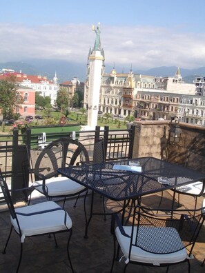 A terrace with a table and chairs, a statue, and a view of a cityscape with buildings and mountains. - Hotel Brighton (Batumi)