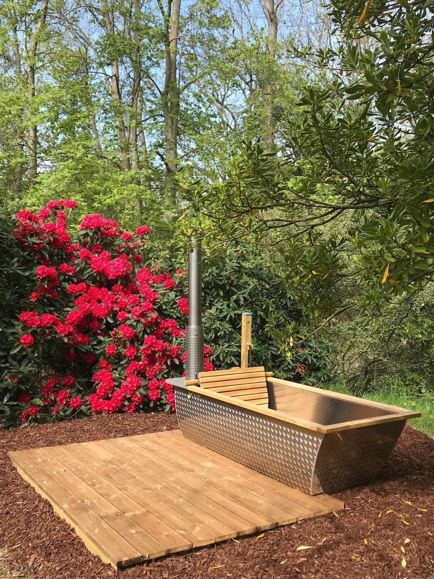 Una terraza de madera con una parrilla de barbacoa incorporada y una chimenea de metal, rodeada de exuberante follaje y flores rojas.