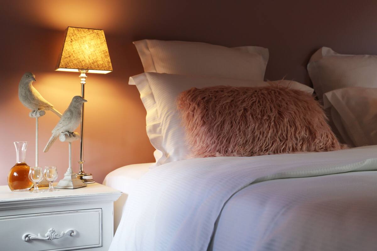A neatly made bed with white bedding and a pink pillow, a bedside table with a lamp, two decorative birds, and a bottle and glasses on the table.