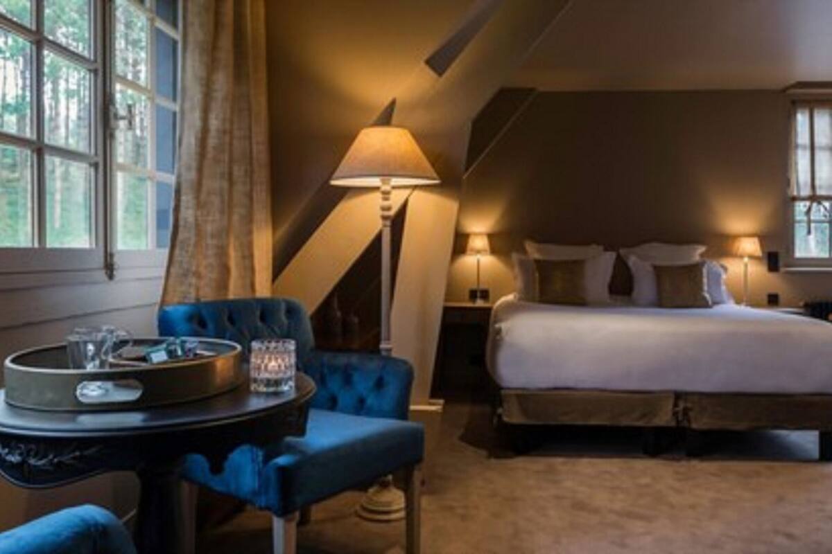 A hotel room with a large bed, a desk with a chair, a lamp, and a view of trees through the window.