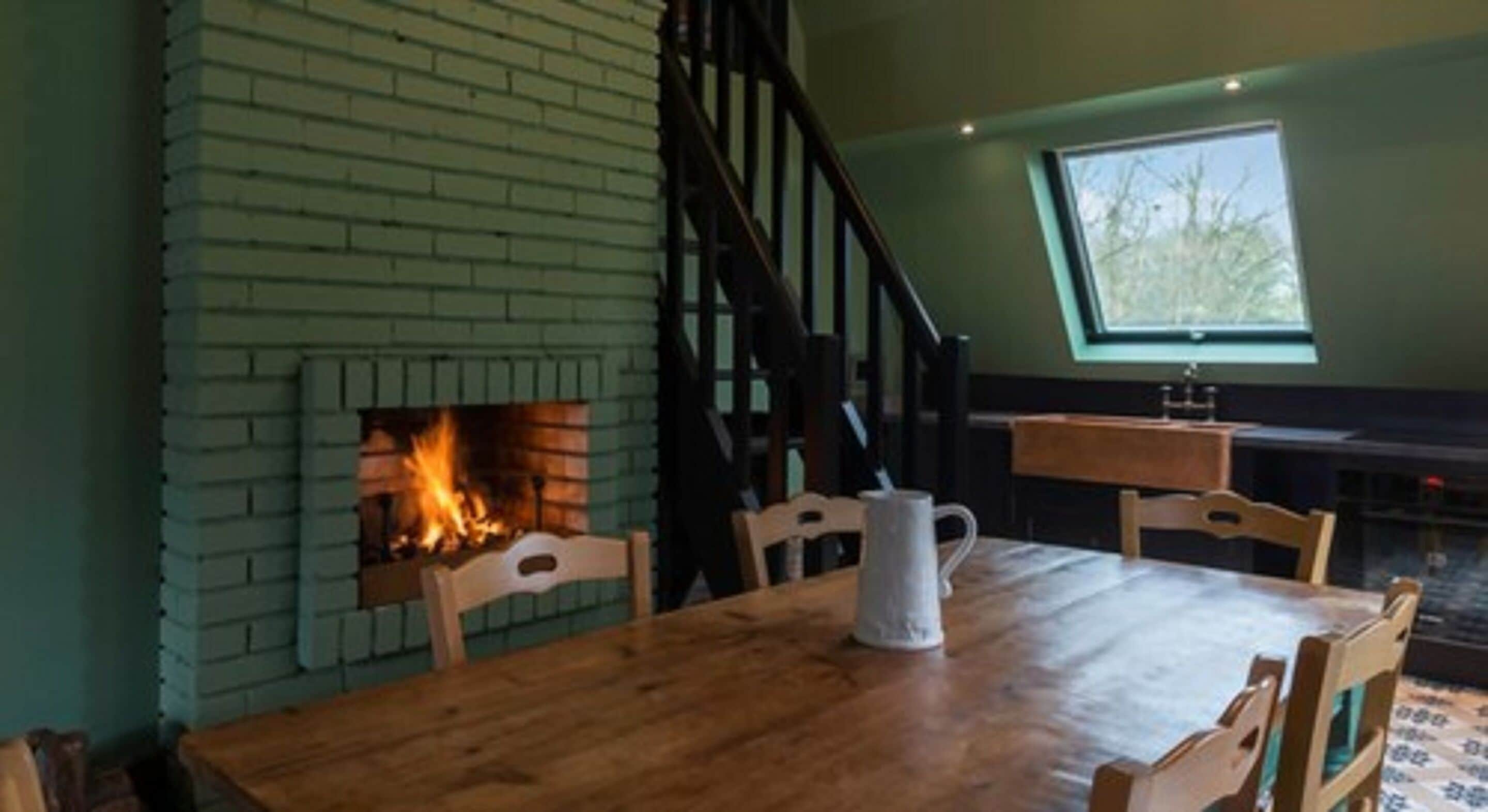 A dining area with a brick fireplace, wooden table, chairs, and a window with a view of trees.