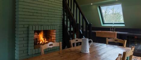 A dining area with a brick fireplace, wooden table, chairs, and a window with a view of trees.