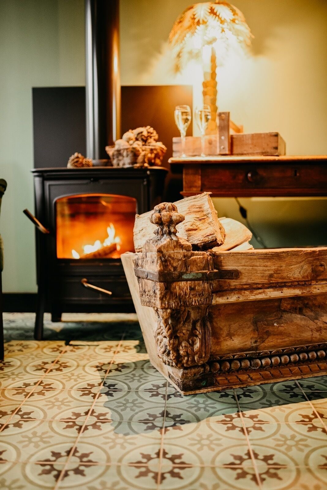 Une pièce chaleureuse avec un poêle à bois, une coffre en bois sculpté et une table avec une lampe.