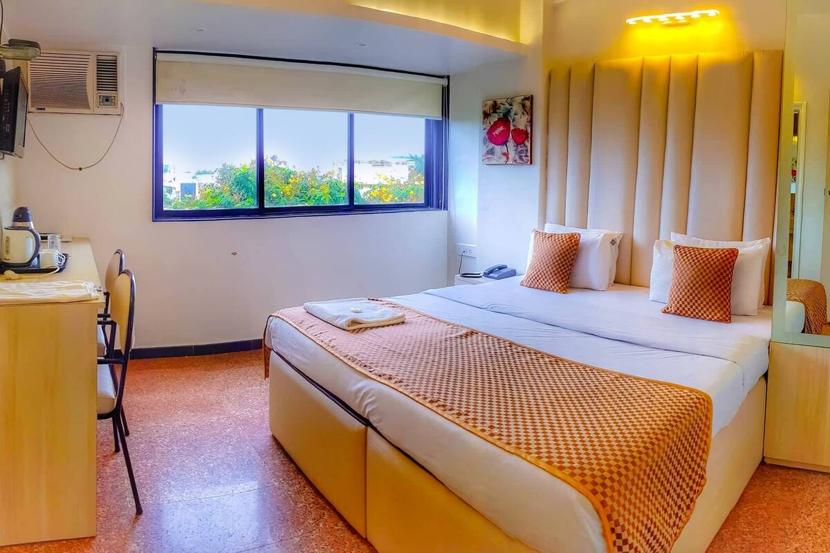 A hotel room with a large bed, a desk with a kettle, and a window with a view of greenery.