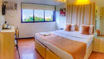 A hotel room with a large bed, a desk with a kettle, and a window with a view of greenery.