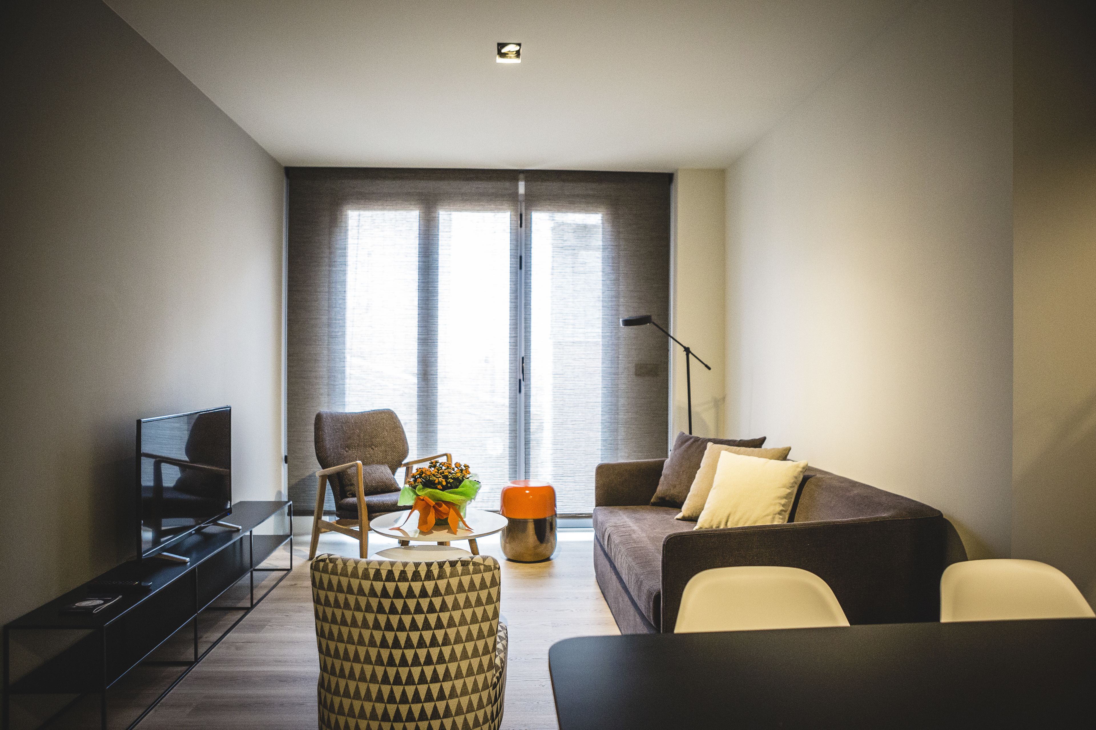 A modern living room with a flat-screen TV, a sofa, a coffee table with a bouquet, and a large window with curtains.