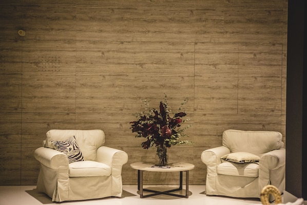 A room with two beige armchairs, a small round table with a vase of flowers, and a wooden wall. - Apartamentos Bárbara 3 (Alicante)