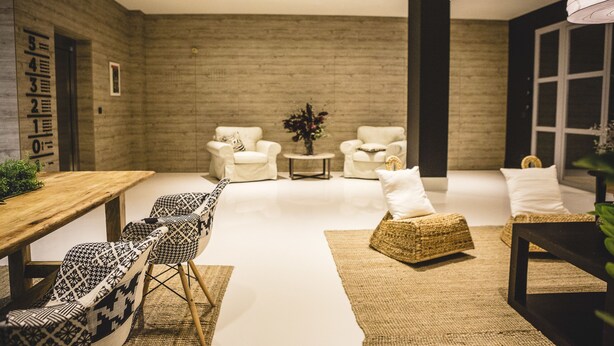 A modern living room with a wooden dining table, patterned chairs, a woven pouf, and a small side table with a plant.