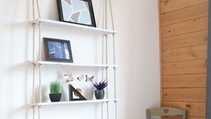 Eine weiße Wand mit einem Makramee-Regal, das Bücher, ein gerahmtes Foto und dekorative Gegenstände trägt. Ein Holzstuhl steht an der Wand.