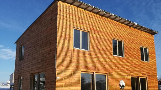 Ein zweistöckiges Holzhaus mit geneigtem Dach, großen Fenstern und einer überdachten Veranda.