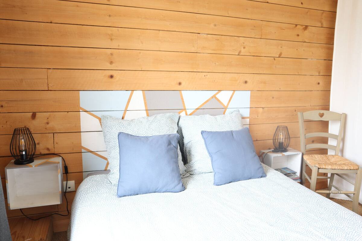 Ein Schlafzimmer mit einem hölzernen Kopfende, einem weißen Nachttisch mit Lampe, einem Sessel und einem Bett mit blauen Kissen.