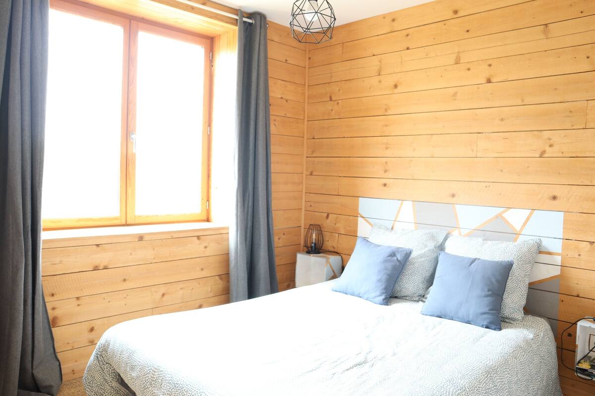 Ein Zimmer mit Holzwänden, einem Bett, einem Nachttisch und einem Fenster mit Vorhängen.