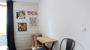 Ein Zimmer mit einem Holztisch, einem schwarzen Stuhl und einem Etagenbett mit einer Holzleiter. An den Wänden befinden sich Wanddekorationen, darunter ein gerahmtes Bild und ein farbenfroher Kunstdruck.