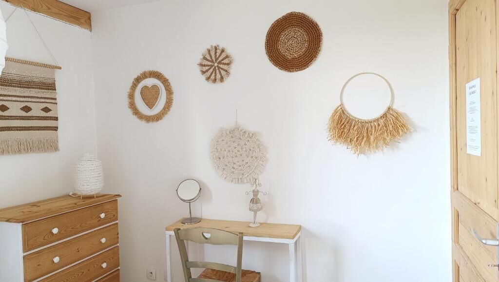 A room with a wooden dresser, a white desk with a chair, and wall decorations including a heart-shaped and a circular woven piece.