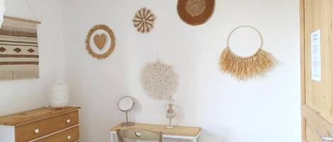 Quarto com cômoda de madeira, escrivaninha branca com cadeira e decorações na parede, incluindo um objeto em formato de coração e uma peça circular trançada.