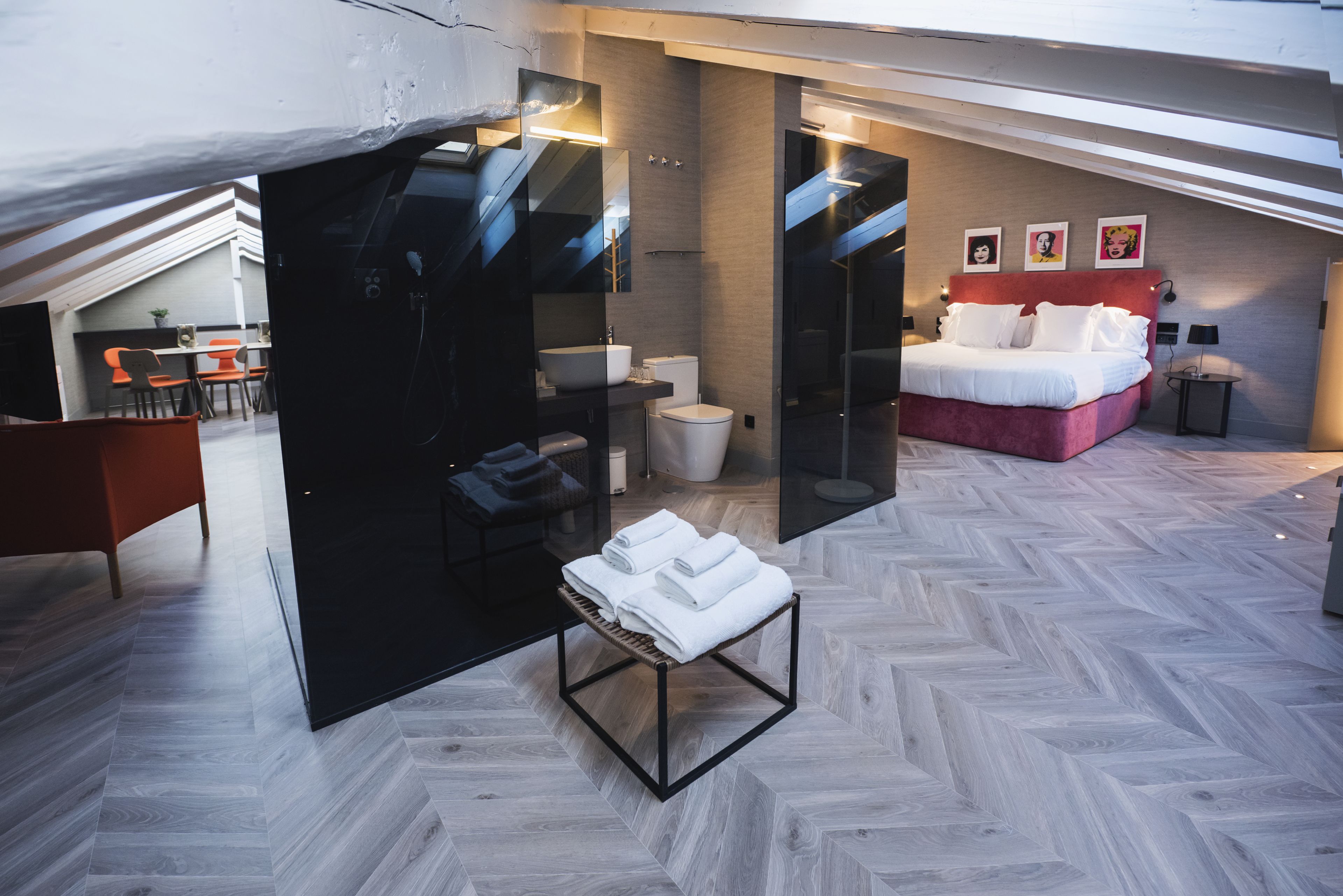 A modern hotel room with a herringbone floor pattern, a bed with white linens, a black-framed bench with towels, a bathroom with a white sink and toilet, and a dining area with orange chairs.