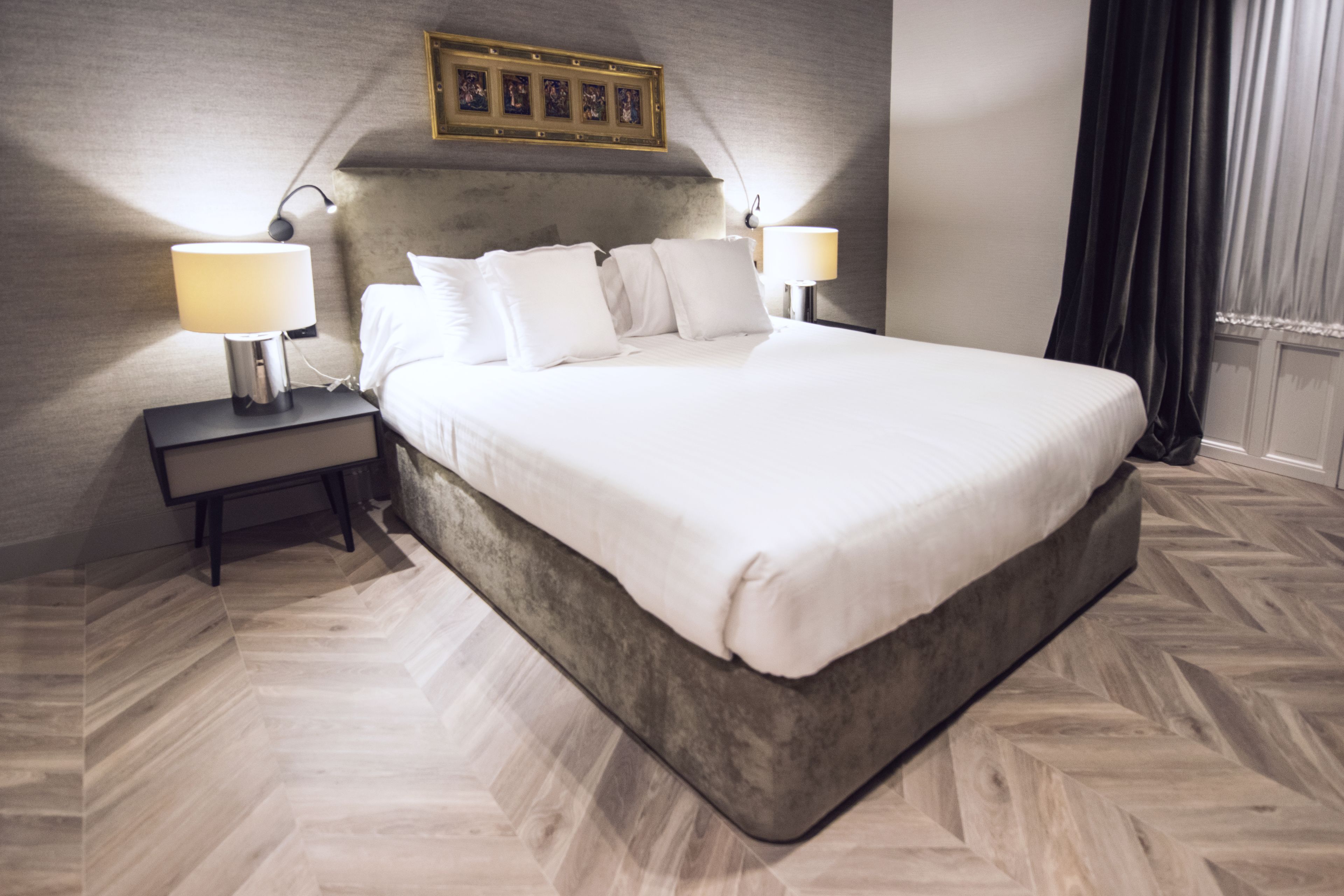 A hotel room with a large bed, two bedside tables with lamps, a headboard with a picture frame, and a herringbone-patterned floor.