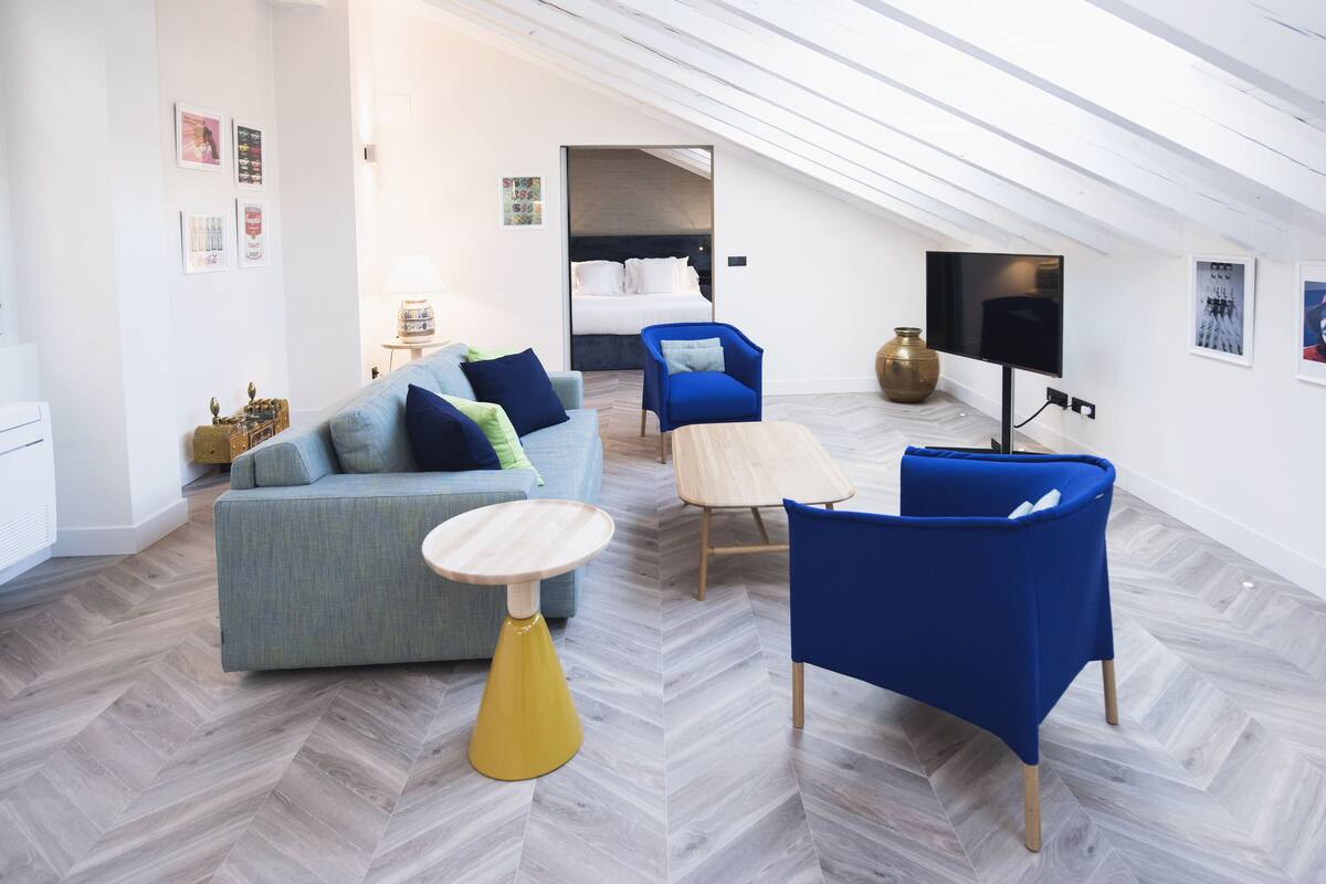A modern, well-lit room with a loft-style ceiling, a grey sofa with blue cushions, a blue armchair, a small wooden table, and a television on the wall.