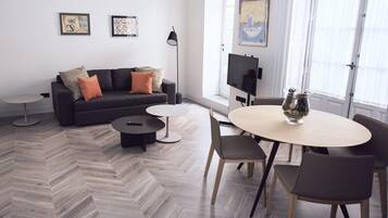 Una sala de estar moderna con un sofá, una mesa de centro y una mesa de comedor. Hay obras de arte enmarcadas en la pared, un televisor y una ventana con persianas.