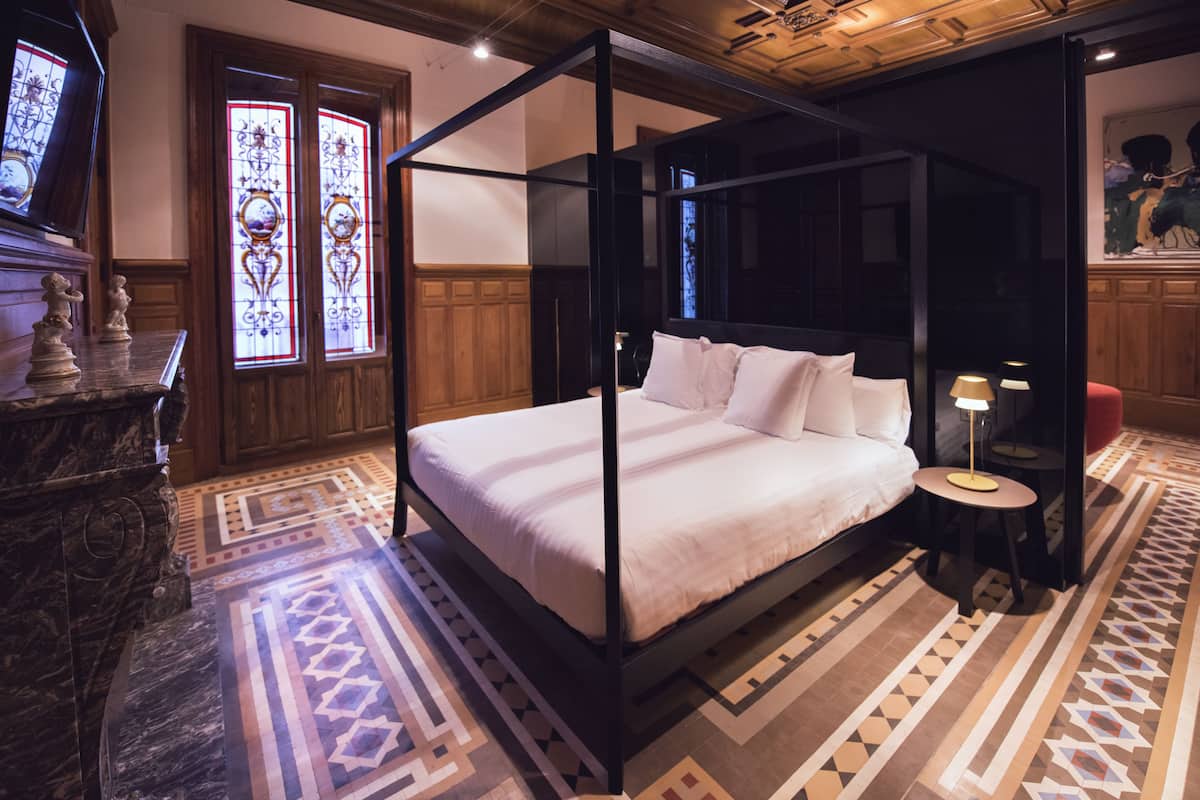 A four-poster bed with white linens, a marble side table, and a patterned floor.
