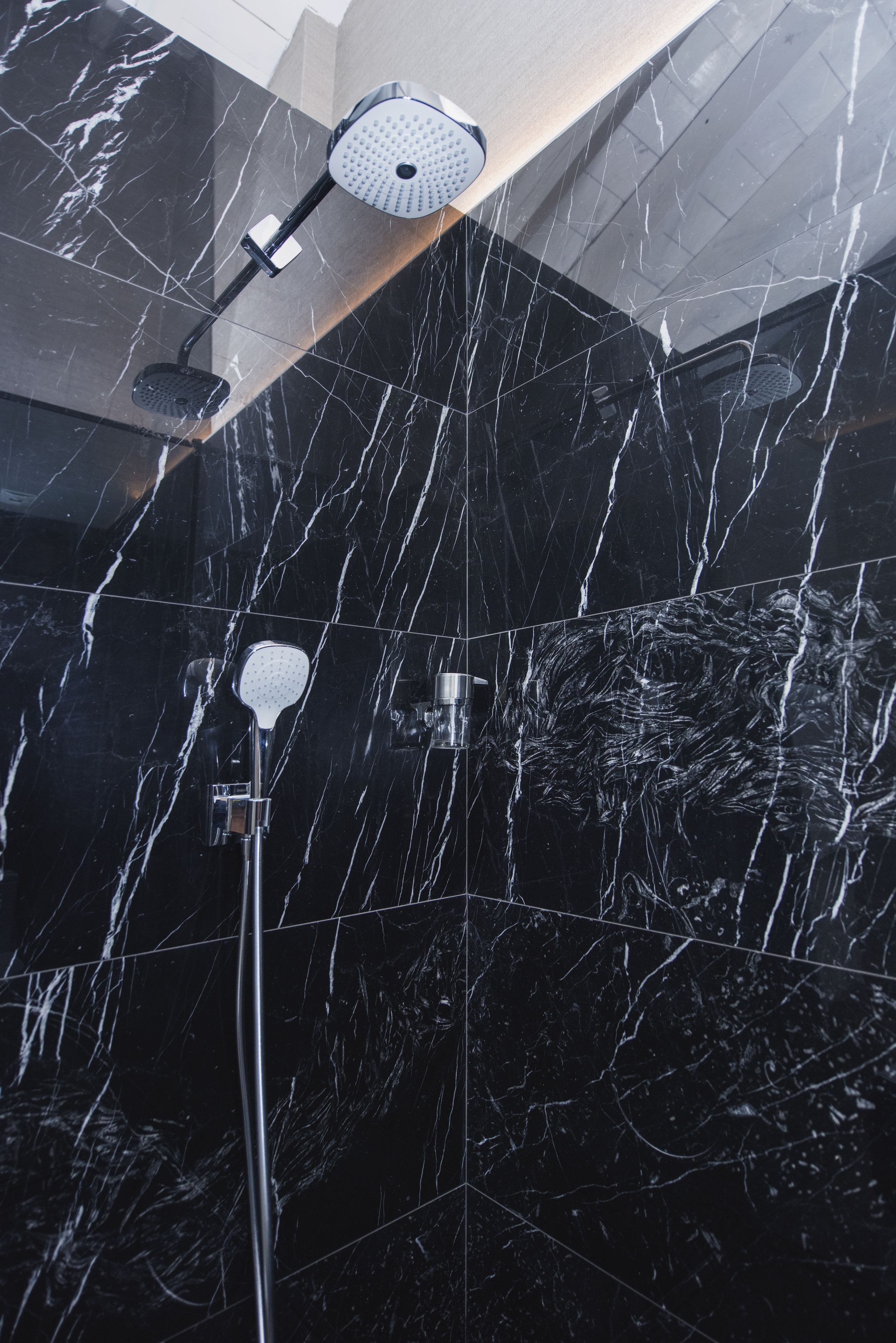 Ducha con cabezal de ducha manual y cabezal de ducha empotrado en un baño con azulejos de mármol negro.