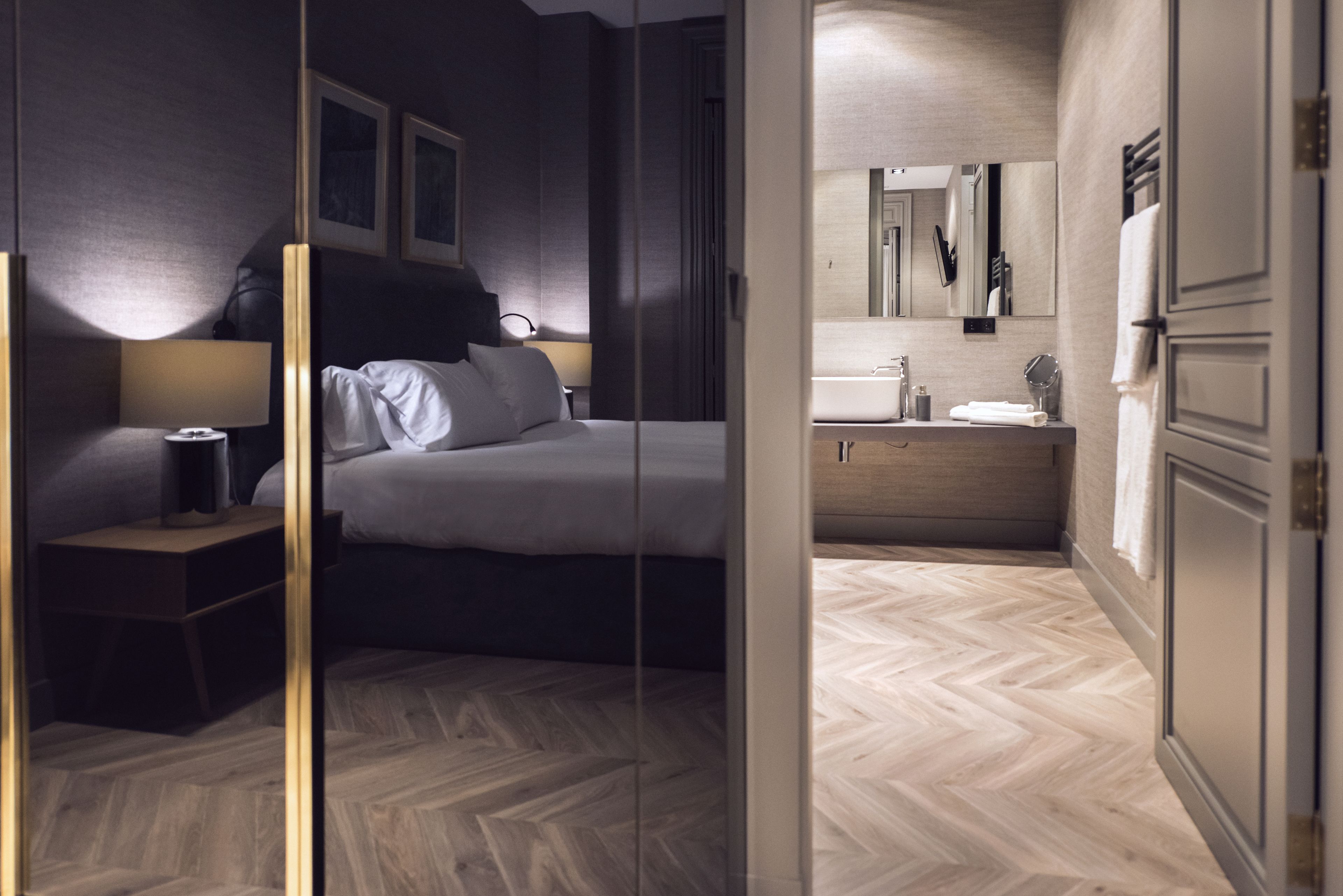 A modern hotel room with a large bed, bedside lamps, a nightstand, a bathroom with a mirror, sink, and towel rack.