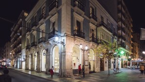 Un bâtiment historique doté d’arches et de balcons, un panneau en forme de croix verte indiquant un établissement médical, et une rue pavée au premier plan.
