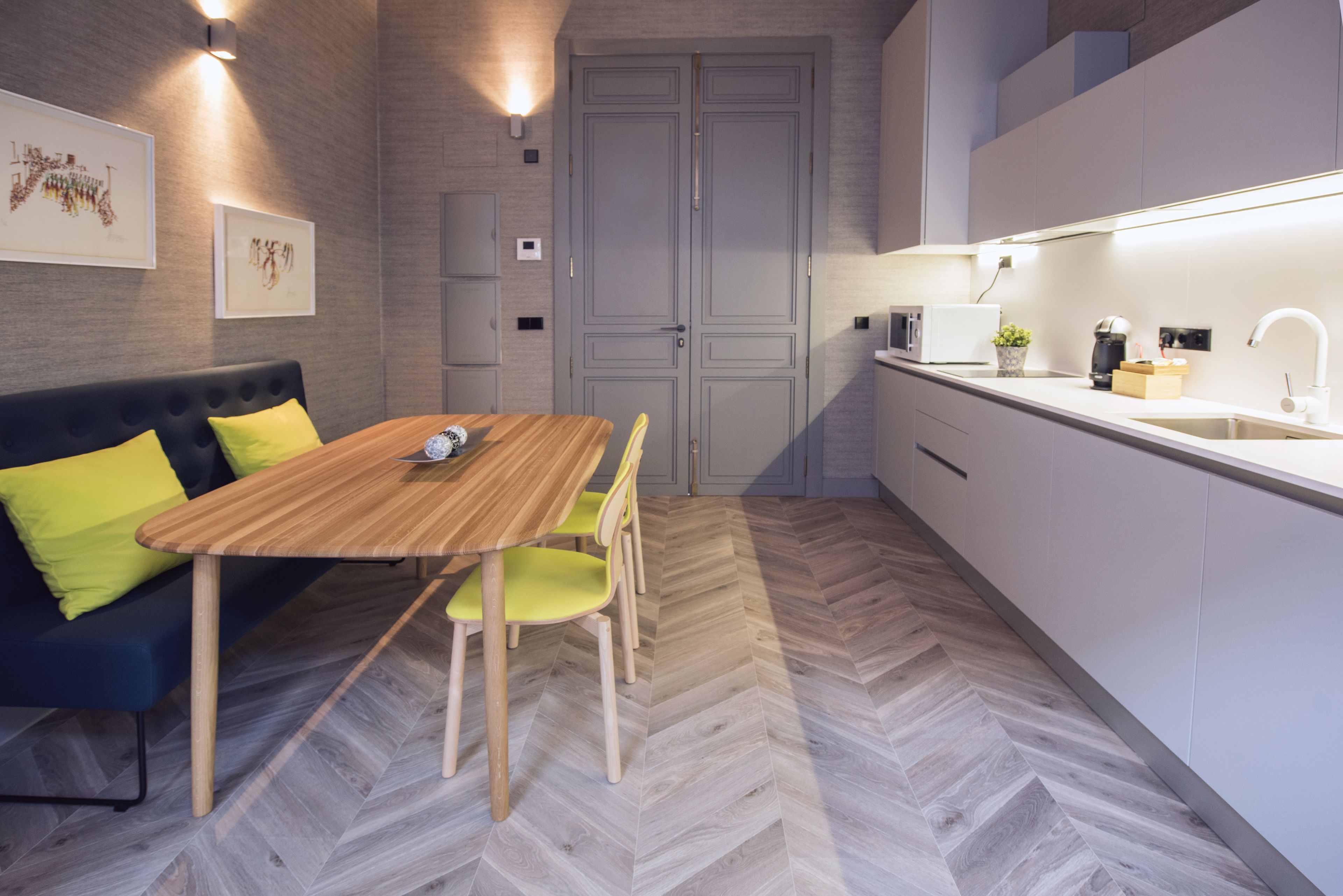 Une cuisine moderne avec un sol en parquet disposé en chevrons, une table à manger en bois avec des chaises, un four encastré et un plan de travail blanc.