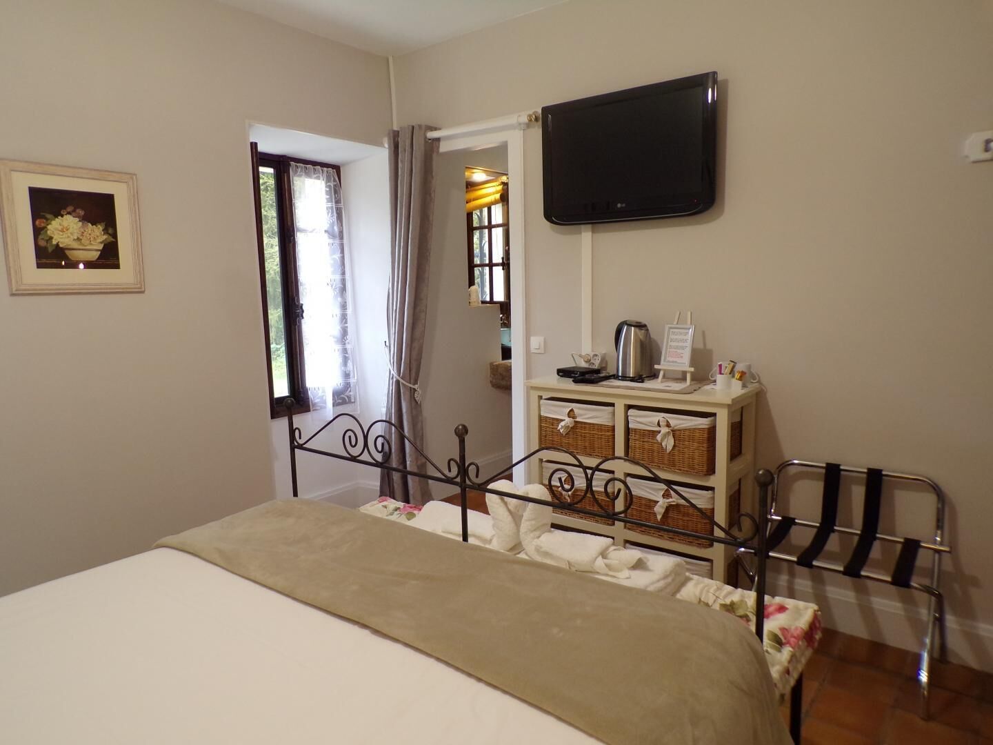 A bedroom with a bed, a television mounted on the wall, a window with curtains, a small table with a kettle and a sign, and a chair with a towel.