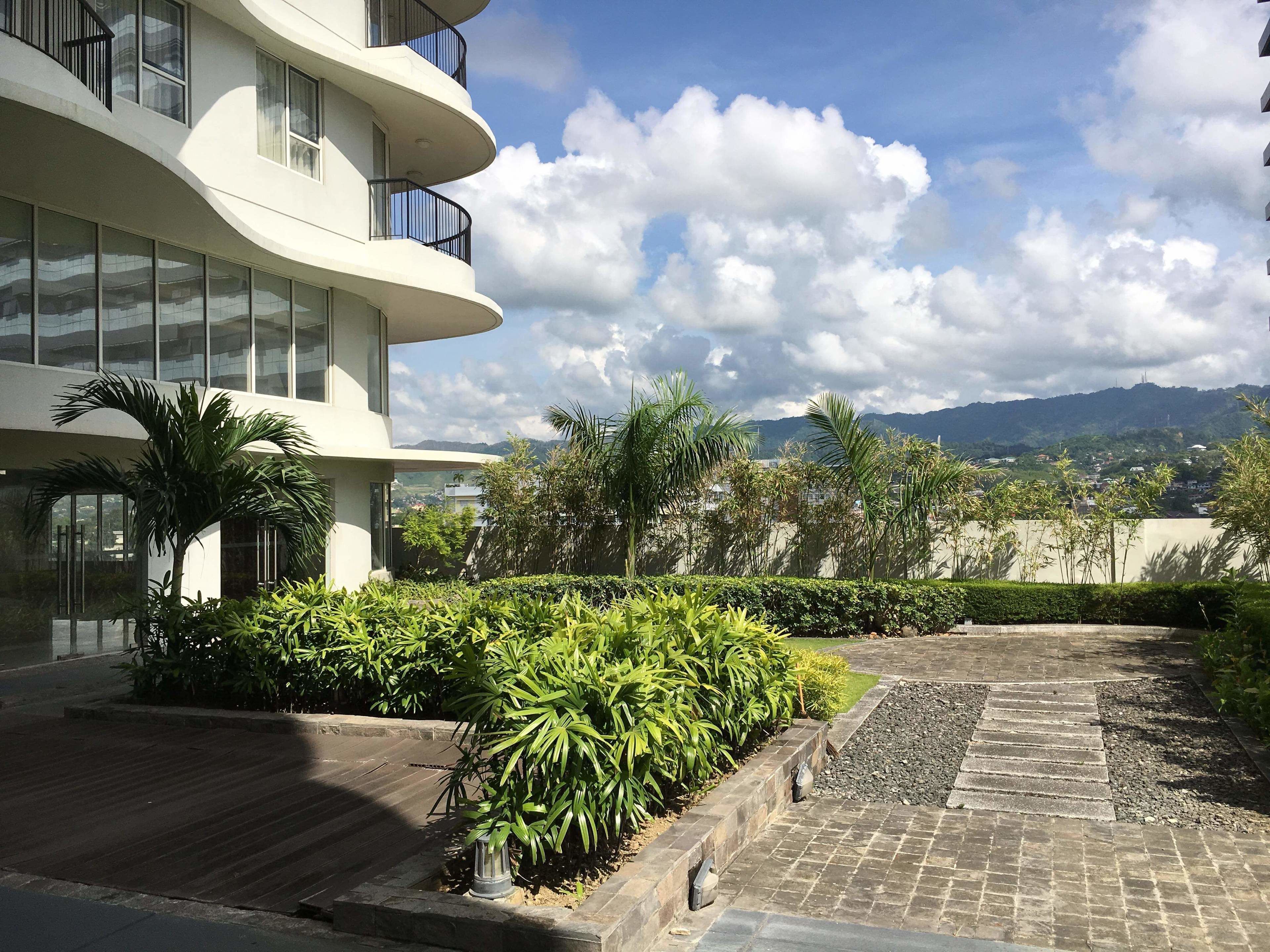 Un edificio moderno con fachada curva, ventanas amplias y un patio ajardinado con palmeras y arbustos.
