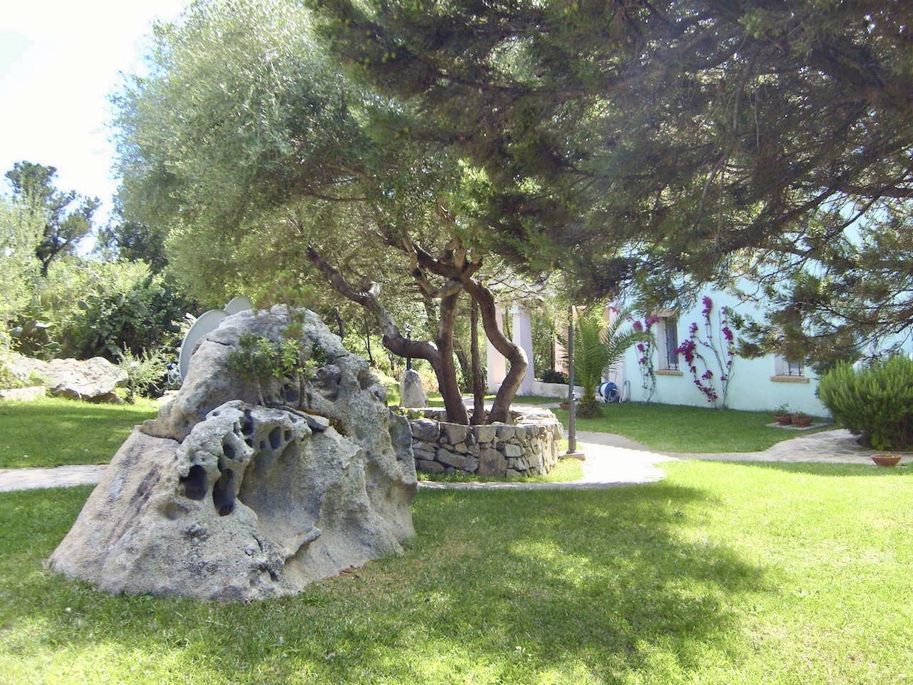 Un jardín con una gran formación rocosa, un árbol y un edificio blanco con una ventana y flores púrpuras.