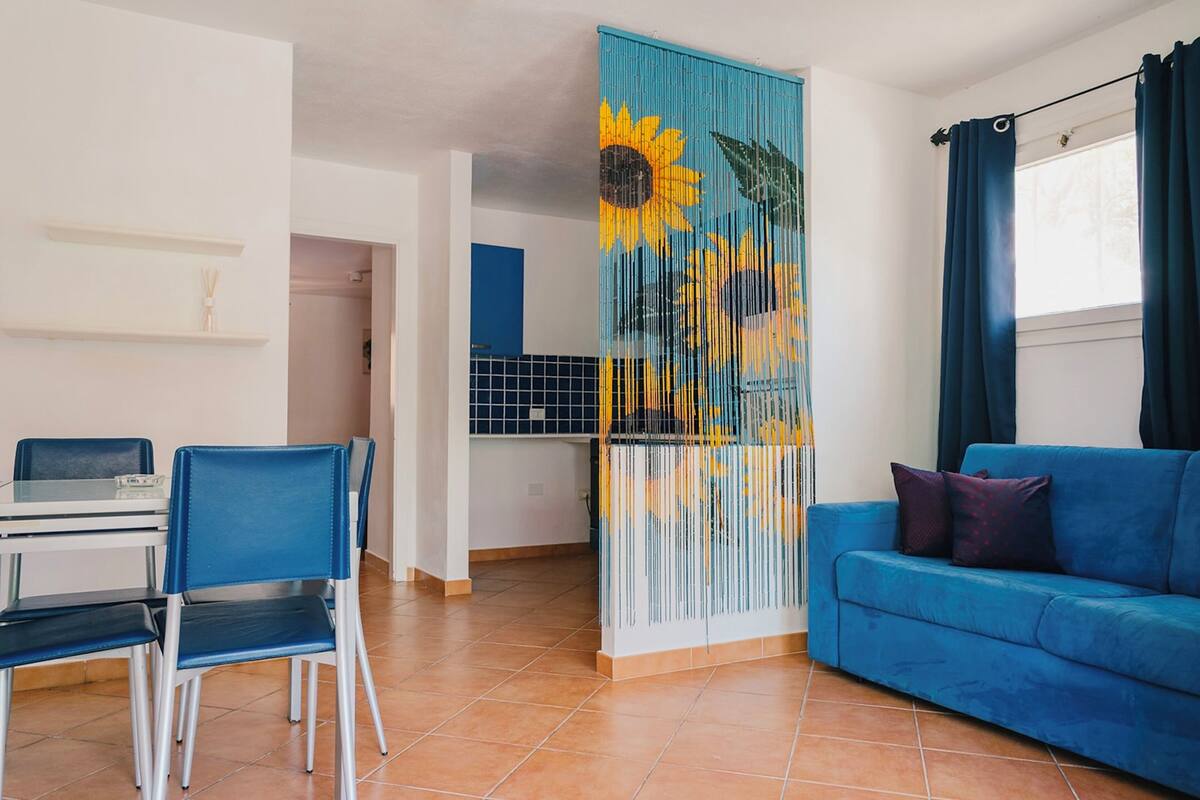 Un salón con un sofá azul, una mesa de comedor con sillas y una pared decorada con motivos de girasoles.
