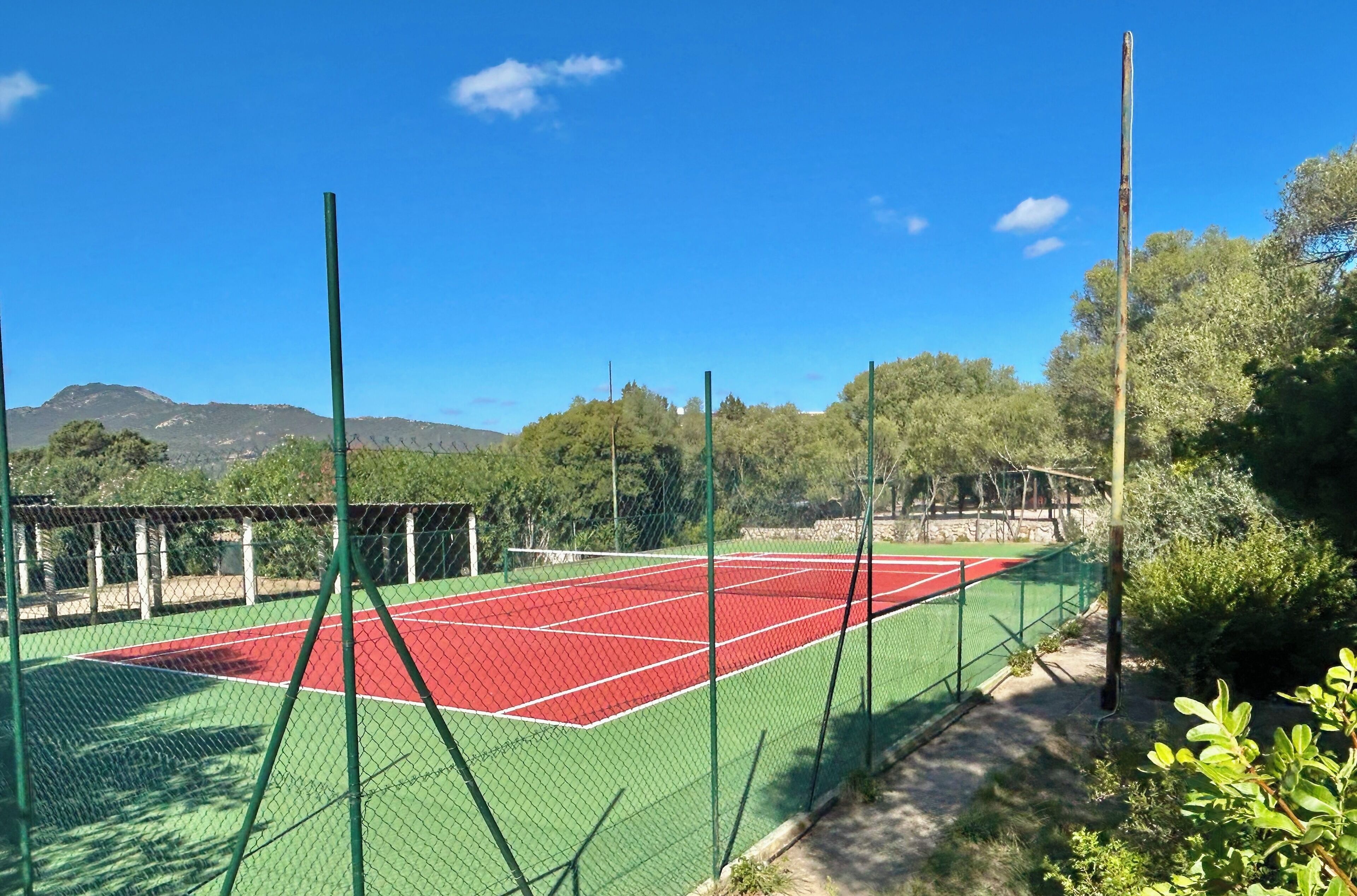 Una cancha de tenis con valla verde, rodeada de árboles y montañas al fondo.