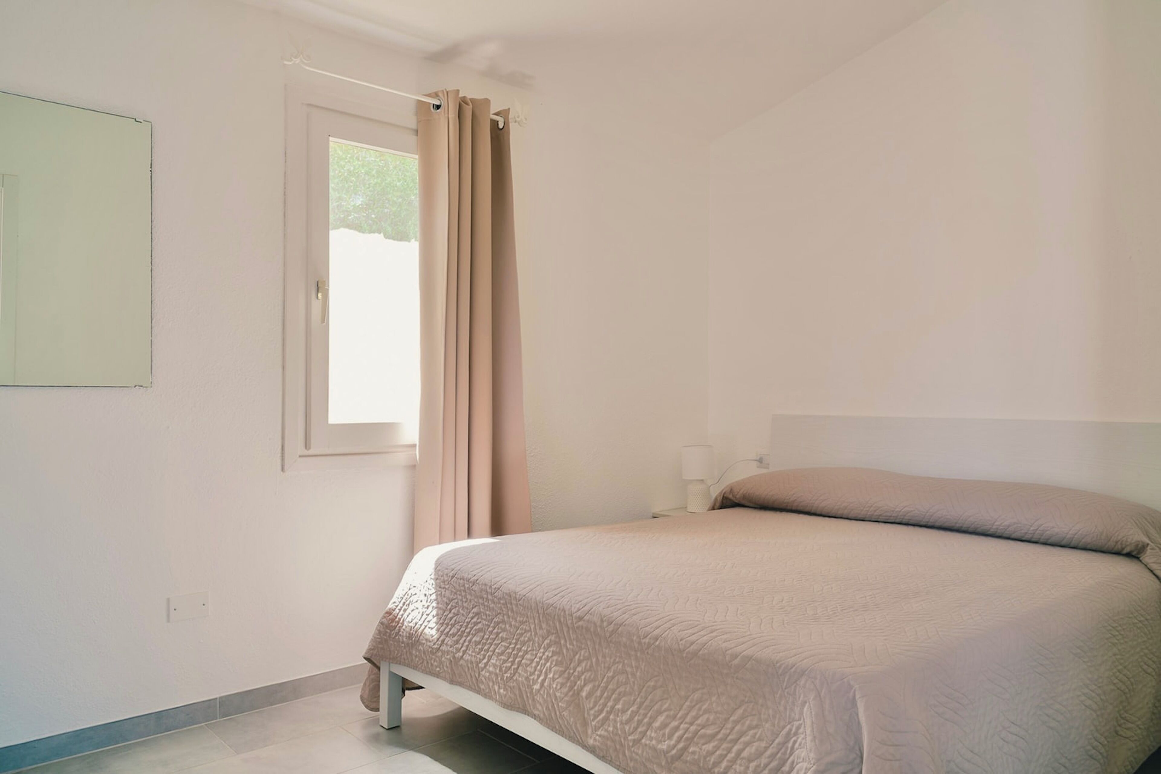 A bedroom with a bed, a window with curtains, a wall-mounted light, and a door.