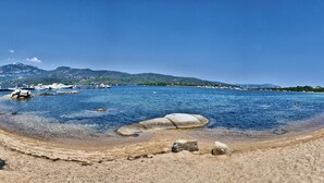 Uma praia costeira com água cristalina, costa rochosa e montanhas ao longe.