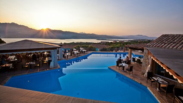 Área junto à piscina com mobiliário externo e vista para o pôr do sol sobre a água.