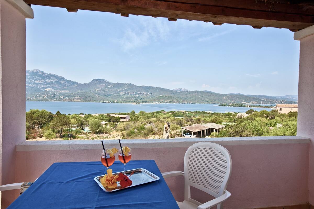Un balcón con una mesa dispuesta para dos personas, con vistas panorámicas a un lago y montañas.