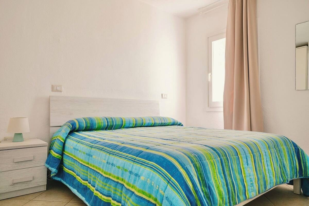 Un dormitorio con una cama, una mesita de noche, una lámpara y una ventana con cortinas.