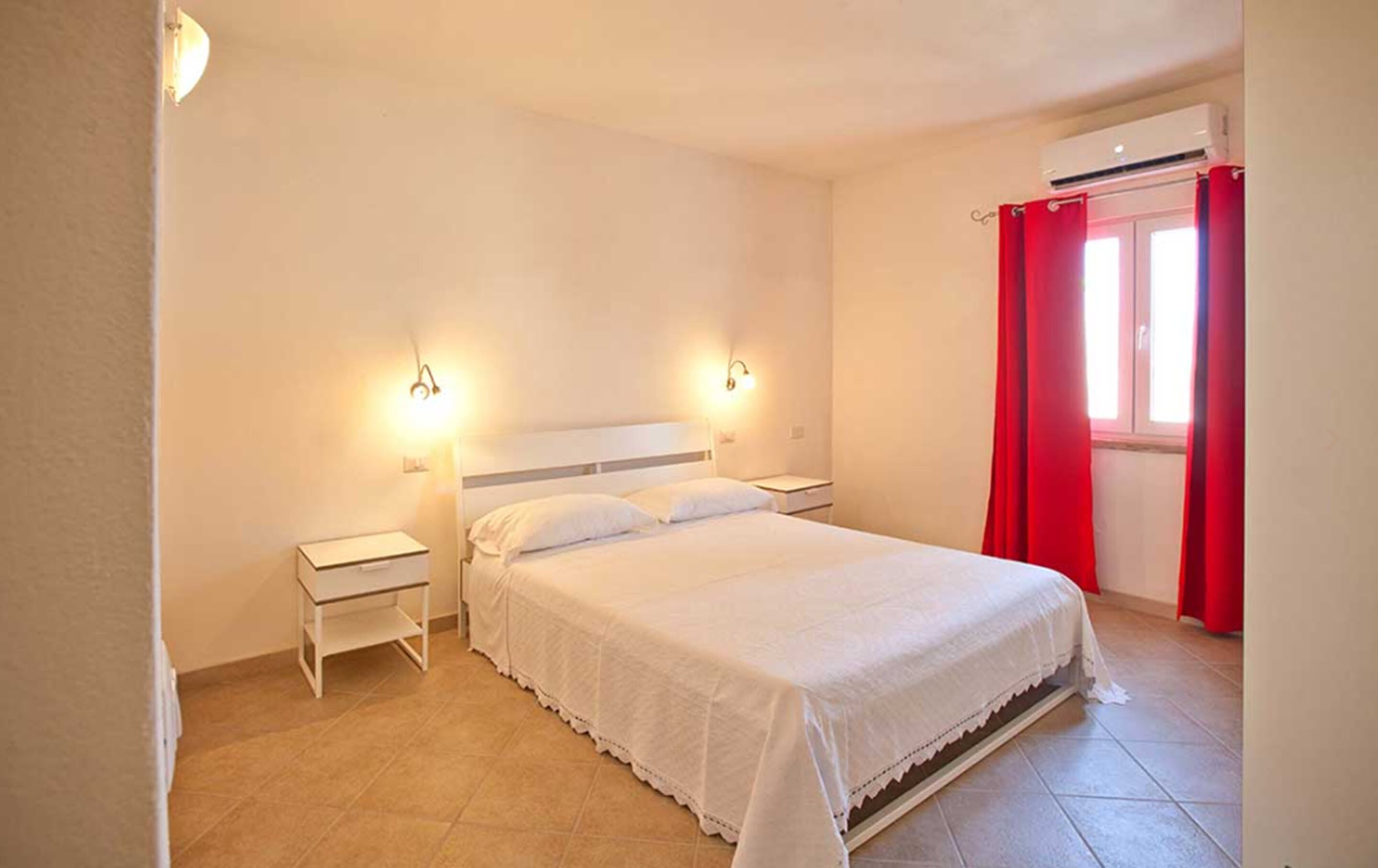 A bedroom with a bed, two bedside tables, a nightstand, a window with red curtains, and an air conditioning unit.