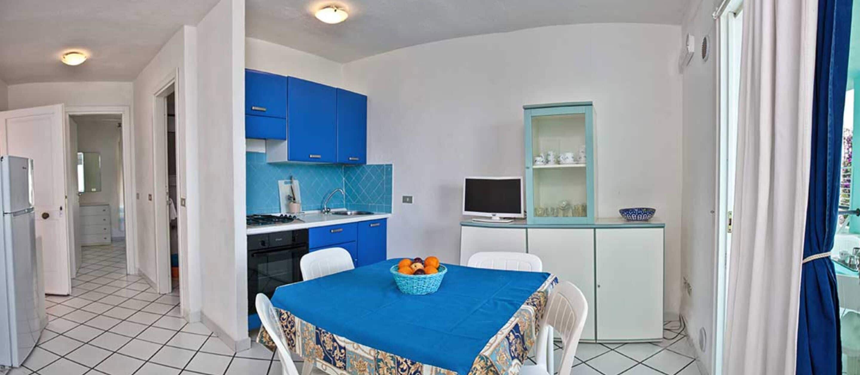 A compact kitchen with blue cabinets, a dining table with a blue tablecloth, and a small TV on a cabinet.