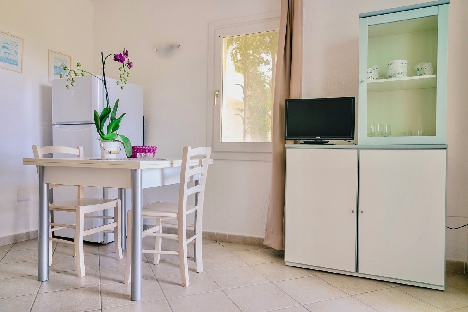 Un comedor con una mesa y sillas, un refrigerador, un televisor sobre un mueble y una ventana con cortinas.