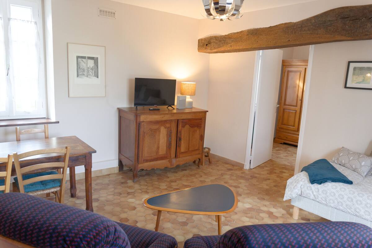 Une pièce comprenant une table à manger en bois, des chaises, un téléviseur posé sur un meuble en bois, un petit tabouret, un lit avec une literie à motifs et un sol carrelé.